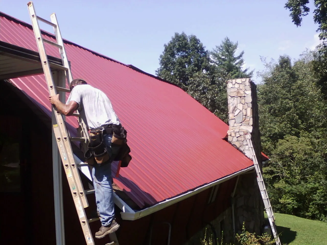 Roofer on a ladder beside a red metal roof and stone chimney — Soffit & Fascia Repair by Roofing Brothers in Milford